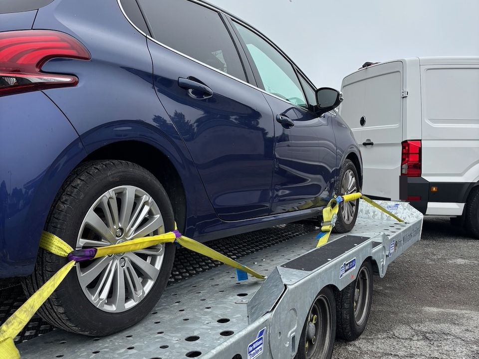 Blue hatchback strapped at the wheels on the car transporter behind the van